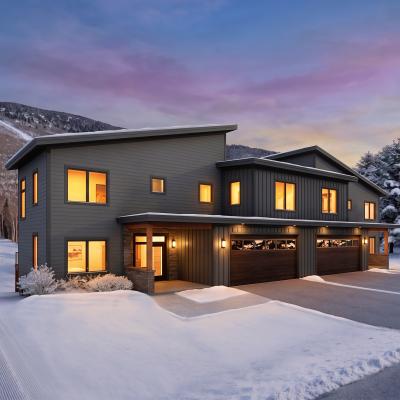 Modern snow-covered house with large windows at dusk, near a skiing trail and pine trees.